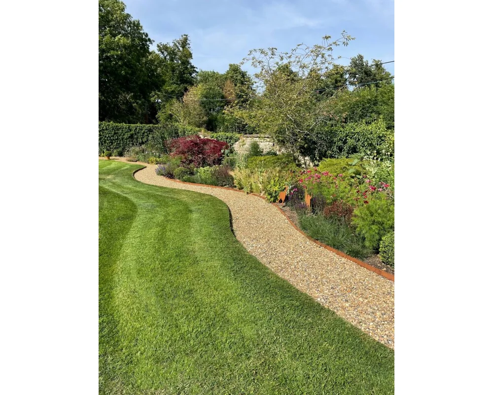 EverEdge PROEDGE Landscape Edging – 4 A winding gravel path, bordered by a well-manicured lawn edging and lush garden beds with colorful plants and shrubs. The background boasts tall trees under a clear blue sky.