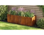 The EverEdge Planter, a rectangular rust-colored metal box, sits against a wooden fence filled with green plants and blooming purple flowers, surrounded by grass and shrubs.