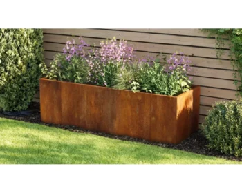 The EverEdge Planter, a rectangular rust-colored metal box, sits against a wooden fence filled with green plants and blooming purple flowers, surrounded by grass and shrubs.